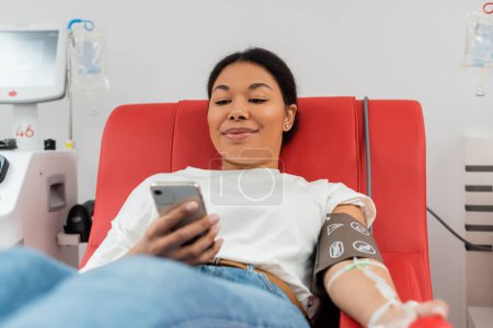 femme multiraciale heureuse dans la messagerie de manchette de pression artérielle sur le téléphone portable alors qu'elle était assise sur une chaise médicale près d'une machine à transfusion automatisée dans un laboratoire moderne, procédure médicale 