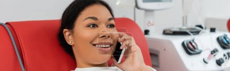 Foto de Mujer multirracial feliz sentado en la silla médica y sonriendo durante la conversación en el teléfono móvil, mientras que la donación de sangre cerca de máquina de transfusión borrosa, pancarta - Imagen libre de derechos
