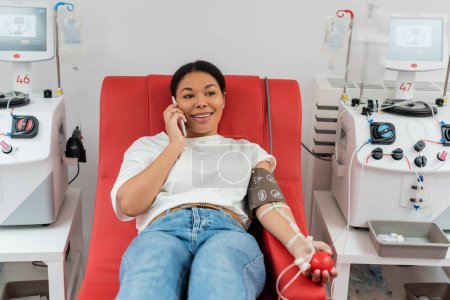Foto de Mujer multirracial sonriente con manguito de presión y bola de goma sentado en la silla médica cerca de las máquinas de transfusión y hablando por teléfono móvil durante la donación de sangre en la clínica - Imagen libre de derechos