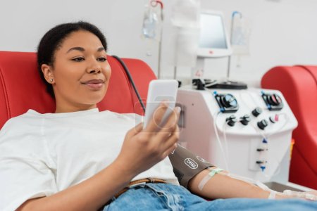 erfreut multirassische Frau sitzt auf bequemen medizinischen Stuhl und Nachrichten auf dem Handy in der Nähe der automatischen Bluttransfusionsmaschine im Labor, verschwommener Hintergrund