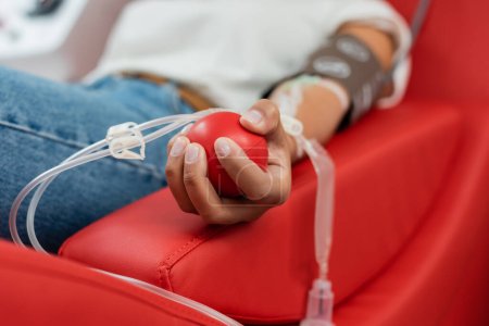 Foto de Enfoque selectivo de la bola de goma en la mano de la mujer multirracial con transfusión conjunto sentado en la silla médica cómoda y la donación de sangre en el laboratorio - Imagen libre de derechos