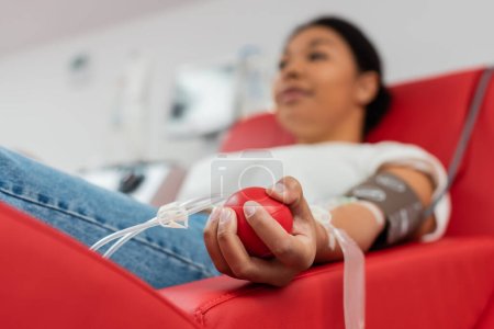 enfoque selectivo de la pelota de goma médica en la mano de la mujer multirracial donando sangre mientras está sentado en la silla médica cómoda en el laboratorio, fondo borroso