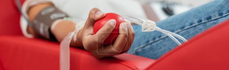 Foto de Vista parcial de la mujer multirracial con el sistema de transfusión que sostiene la bola de goma mientras está sentado en la silla médica ergonómica durante la donación de sangre en el laboratorio, pancarta - Imagen libre de derechos