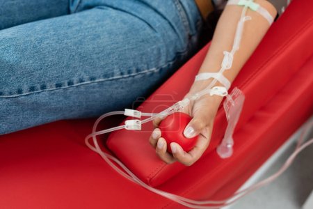 Teilbild einer multirassischen Frau mit einem Transfusionsset, das einen Gummiball hält, während sie während der Blutspende in der Klinik auf einem ergonomischen Behandlungsstuhl sitzt, medizinischer Eingriff 
