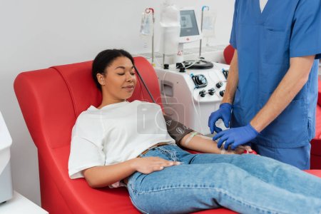 trabajador de la salud en uniforme azul y guantes de látex ajuste transfusión conjunto cerca de la mujer multirracial sentado en la silla médica y la donación de sangre en la clínica, procedimiento médico 