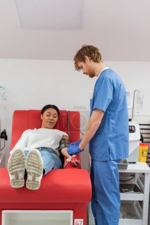 pelirroja trabajadora de la salud en uniforme azul ajustando transfusión fijada en mujer multirracial sentada con pelota de goma en silla médica mientras dona sangre en clínica