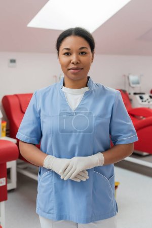 enfermera multirracial positiva en uniforme azul y guantes de látex mirando a la cámara cerca de sillas médicas y máquinas de transfusión en el centro de donación de sangre sobre fondo borroso