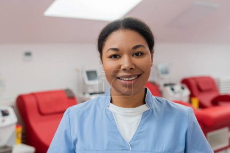 alegre enfermera multirracial en uniforme azul sonriendo y mirando a la cámara cerca de sillas médicas y máquinas de transfusión sobre fondo borroso en el centro de donación de sangre