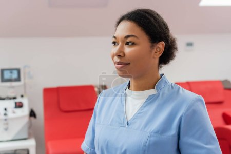 young and pleased multiracial healthcare worker in blue uniform looking away near transfusion machine and medical chairs in blood donation center on blurred background