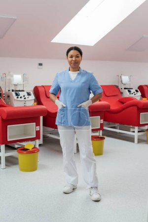 Foto de Enfermera multirracial positiva en guantes de látex de pie con las manos en bolsillos de uniforme cerca de sillas médicas, máquinas de transfusión y cubos de basura en el centro de donación de sangre - Imagen libre de derechos