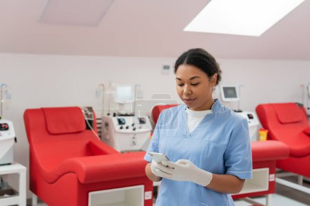 multiracial nurse in blue uniform and latex gloves messaging on mobile phone near medical chairs and transfusion machines in blood donation center
