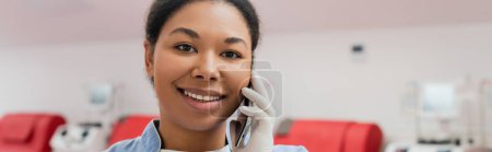 young and happy multiracial healthcare worker in sterile latex glove smiling during conversation on mobile phone in blood transfusion station, banner