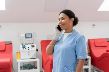 trabajador sanitario multirracial feliz en uniforme azul y guante de látex hablando en el teléfono móvil cerca de la máquina de transfusión borrosa y sillas médicas en el centro de donación de sangre