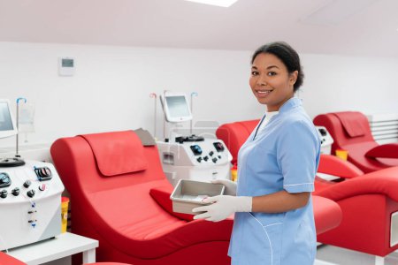 Trabajador sanitario multirracial en uniforme azul y guantes de látex sosteniendo bandeja médica cerca de máquinas de transfusión y cómodas sillas médicas en el centro de donación de sangre