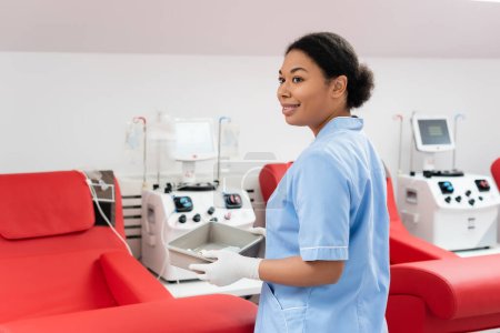 Lächelnde Krankenschwester in blauer Uniform und Latexhandschuhen mit medizinischem Tablett in der Nähe von Transfusionsmaschinen und ergonomischen Stühlen im Blutspendezentrum