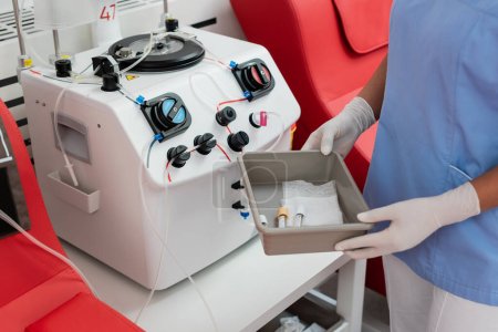 vista parcial del trabajador sanitario multirracial en guantes uniformes y de látex sosteniendo bandeja médica con tubos de ensayo cerca de la máquina de transfusión en el centro de donación de sangre