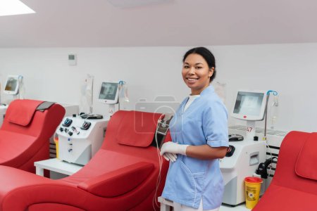alegre enfermera multirracial en uniforme azul y guantes de látex mirando a la cámara cerca de sillas médicas y máquinas de transfusión con monitores en el centro de donación de sangre