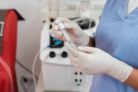 Teilansicht des multirassischen Gesundheitspersonals in blauer Uniform und Latexhandschuhen mit Reagenzglas in der Nähe eines automatisierten Transfusionsgeräts auf verschwommenem Hintergrund in der Klinik