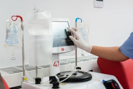Téléchargez les photos : Vue recadrée de l'infirmière multiraciale en gant de latex faisant fonctionner la machine de transfusion avec écran tactile près des stands de goutte à goutte avec des poches de perfusion dans le centre de don de sang - en image libre de droit