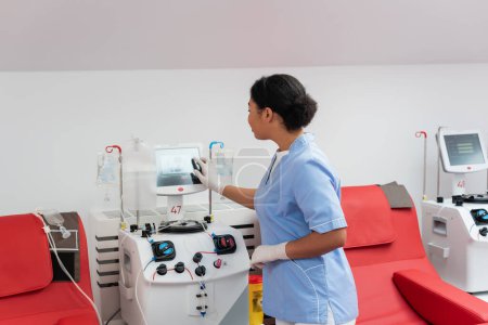 joven médico multirracial en uniforme azul y guantes de látex operando máquina de transfusión con pantalla táctil cerca de sillas médicas en el centro de donación de sangre
