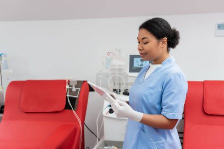 positive multirassische Gesundheitshelferin in blauer Uniform und Latexhandschuhen mit digitalem Tablet in der Nähe von Medizinstühlen und Transfusionsmaschine im Blutspendezentrum