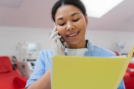 fröhliche multiethnische Krankenschwester in blauer Uniform und Latex-Handschuh, die auf dem Smartphone spricht und in der Nähe verschwommener Transfusionsmaschinen und medizinischer Stühle im Blutspendezentrum auf Papiermappen blickt