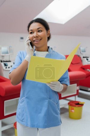 Lächelnde Krankenschwester in blauer Uniform und Latex-Handschuhen mit Papiermappe und Handy in der Nähe verschwommener medizinischer Stühle und Transfusionsmaschinen im Blutspendezentrum