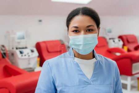 Photo for Young multiracial nurse in blue uniform and medical mask looking at camera near medical chairs and transfusion machines on blurred background in hospital - Royalty Free Image