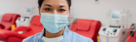 young multiracial healthcare worker in blue uniform and medical mask looking at camera near blurred medical chairs and transfusion machines in blood donation center, banner