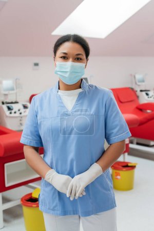 enfermera multirracial en uniforme azul, máscara médica y guantes de látex mirando a la cámara cerca de las máquinas de transfusión, sillas médicas y cubos de basura sobre fondo borroso en el centro de donación de sangre