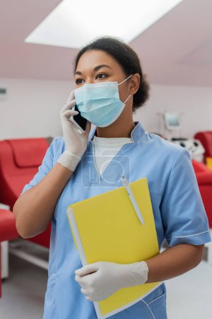 multiracial nurse in blue uniform, medical mask and latex gloves holding paper folder and pen while talking on smartphone near blurred medical chairs in blood transfusion station