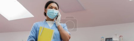 low angle view of multiracial nurse in blue uniform, medical mask and latex glove holding paper folder with pen and talking on mobile phone in blood donation center, banner