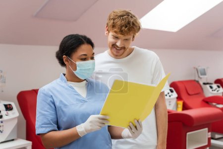 Krankenschwester in blauer Uniform, medizinischer Maske und Latexhandschuhen zeigt lächelnden rothaarigen Mann Papiermappe in der Nähe von Transfusionsmaschinen und verschwommenen Medizinstühlen im Blutspendezentrum