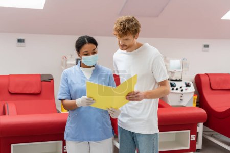 redhead volunteer and multiracial nurse in uniform, medical mask and latex gloves looking at paper folder near medical chairs and transfusion machines in blood donation center