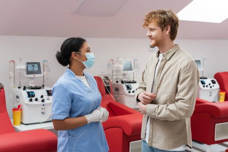 multiracial nurse in blue uniform, medical mask and latex gloves talking to redhead volunteer near transfusion machines and medical chairs in laboratory