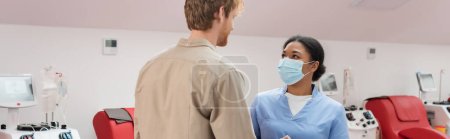 young redhead blood donor and multiracial nurse in blue uniform and medical mask talking near medical chairs and transfusion machines in hospital, banner