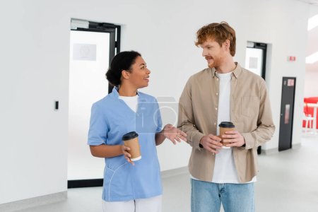 young redhead volunteer and smiling multiracial healthcare worker in uniform holding paper cups while talking in waiting area of blood donation center 