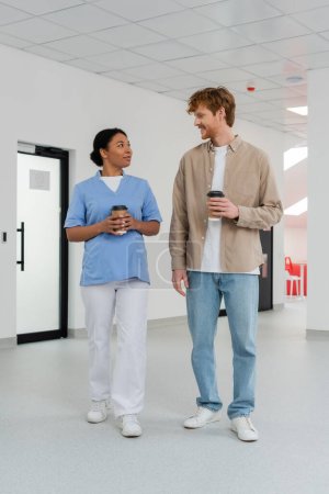 multiracial nurse in uniform and young, positive redhead volunteer holding paper cups with coffee and talking in waiting area of blood donation center, door on background 