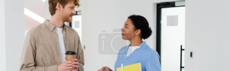 Photo for Young and positive redhead man with takeaway coffee and multiracial nurse with paper folder and pen talking in corridor of blood donation service, banner, door on background - Royalty Free Image