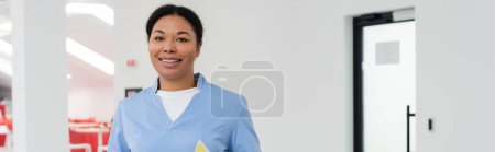 fröhliches multiethnisches Gesundheitspersonal in blauer Uniform, das in die Kamera blickt, während es im Wartebereich eines modernen Blutspendezentrums steht, Banner, Tür im Hintergrund 