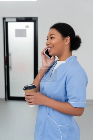 fröhliche multiethnische Gesundheitshelferin in blauer Uniform, Pappbecher mit Kaffee zum Mitnehmen in der Hand und lächelnd während des Gesprächs auf dem Handy im Saal des Blutspendezentrums, Tür im Hintergrund 
