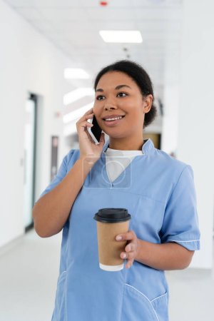 positive multirassische Krankenschwester lächelt während eines Gesprächs auf dem Smartphone, während sie mit Kaffee zum Mitnehmen im Flur der Bluttransfusionsstation steht, verschwommene Halle im Hintergrund 