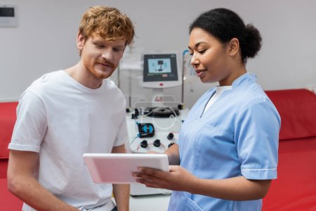 Mitarbeiter des multiethnischen Gesundheitswesens zeigt lächelnden rothaarigen Mann ein digitales Tablet in der Nähe eines verschwommenen Transfusionsgeräts und medizinischer Stühle im Blutspendezentrum