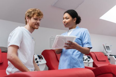 Krankenschwester in blauer Uniform zeigt einem glücklichen rothaarigen Blutspender ein digitales Tablet, der auf einem bequemen medizinischen Stuhl in der Nähe eines automatisierten Transfusionsautomaten im Krankenhaus sitzt