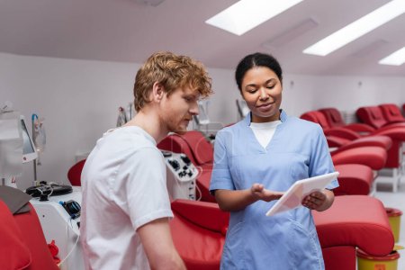 positive multirassische Krankenschwester in blauer Uniform zeigt auf digitales Tablet in der Nähe junger rothaariger Mann und bequeme medizinische Stühle mit Transfusionsmaschinen in Blutspendezentrum