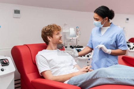 Téléchargez les photos : Infirmière multiraciale en masque médical et gants de latex touchant l'épaule et parlant au donneur de sang roux sur la chaise médicale près de la machine de transfusion dans la clinique - en image libre de droit