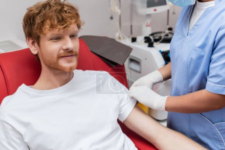 donante de sangre pelirroja positivo sonriendo cerca de enfermera multirracial en uniforme azul y guantes de látex mientras está sentado en la silla médica junto a la máquina de transfusión borrosa en el centro de donación de sangre