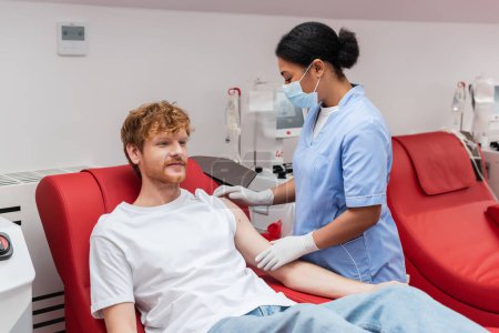multirassische Krankenschwester in medizinischer Maske und Latexhandschuhen, die den Arm eines rothaarigen Blutspenders betrachtet, der auf einem bequemen medizinischen Stuhl in der Nähe einer Transfusionsmaschine im Krankenhaus sitzt