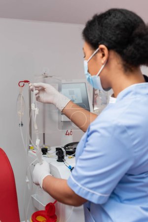 multiracial nurse in blue uniform and latex gloves holding blood transfusion set near drip stand with infusion bags in laboratory, blurred foreground