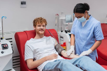 redhead volunteer sitting on medical chair near automated equipment while multiracial nurse in medical mask fixing transfusion set in blood donation center
